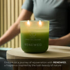 Green candle labeled 'Renewed' on a wooden surface with a blurred person in the background.