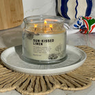 Sun-Kissed Linen 3-wick jar candle on a decorative plate with a woven mat and kitchen items in the background.