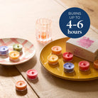 Colorful tealight candles on a table with a box and text indicating burn time.