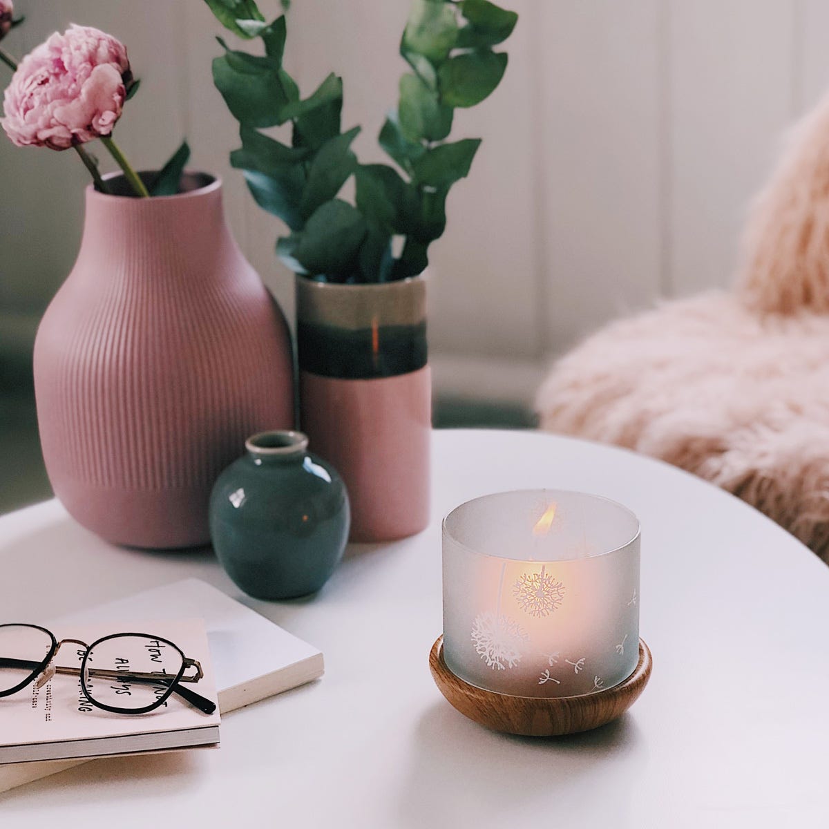 Springtime Blossoms Large Tealight Holder with a wooden base and a frosted glass insert with etched blossoms, shown with a lit tealight on a white table with vases of glasses and books with a pair of glasses on top of.