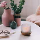 Springtime Blossoms Large Tealight Holder with a wooden base and a frosted glass insert with etched blossoms, shown with a lit tealight on a white table with vases of glasses and books with a pair of glasses on top of.